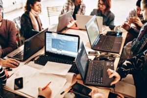 A group of people collaborating around a table full of laptops, papers, and notebooks. They seem engaged in a project or discussion, with one laptop displaying design software. The atmosphere is casual and focused.