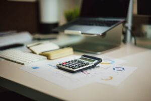 A calculator sitting on an office desk alongside some papers showing graphs and a laptop setup.