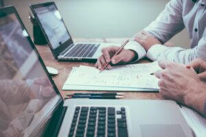 The forearms of two men in shirts planning on A4 paper next to their laptops on a wooden desk.